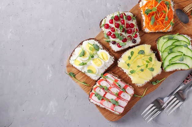 Seis sándwiches en tostadas con zanahorias frescas, pepinos, piña, grosella roja, palitos de cangrejo y huevos de codorniz con microgreens de guisantes sobre tabla de madera sobre fondo gris, vista desde arriba, espacio de copia