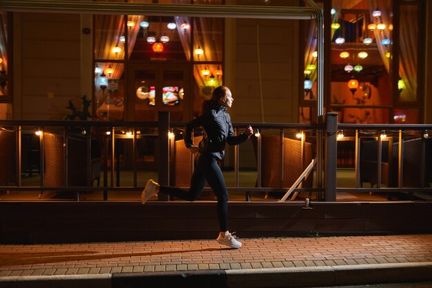 Seitenansicht auf junge kaukasische Sportlerin, die beim Nachtsport im Freien am Stadtcafé vorbeiläuft, konzentrierte sich auf...