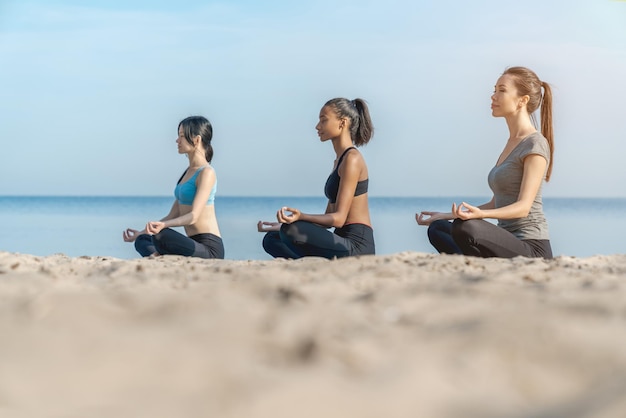 Seitenansicht Aufnahme von Frauen, die Yoga-Meditation in Lotus-Pose am sonnigen Strand in der Nähe von Wasser machen