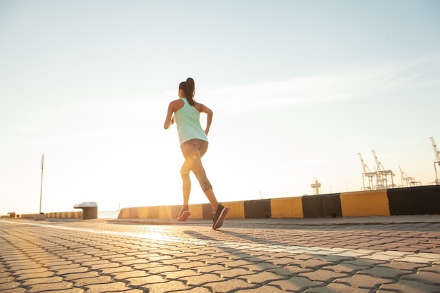 Seitenansicht der Fitnessfrau, die auf einer Straße am Meer in der Nähe des Seehafens läuft. Sportlertraining auf der Strandpromenade bei Sonnenuntergang.