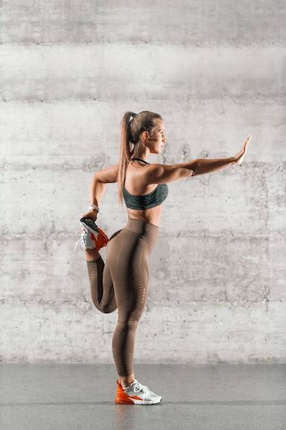 Seitenansicht der flexiblen Brünette mit Pferdeschwanz und in der Sportbekleidung, die Bein vor grauer Wand im Fitnessstudio streckt.