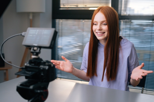 Seitenansicht der fröhlichen jungen Bloggerin schaltet die Kamera ein, winkt mit der Hand und beginnt zu sprechen, um in die Kamera zu schauen. Lächelnder rothaariger weiblicher Rekord kommerzieller Online-Videokurs im Homeoffice.