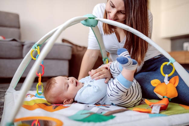 Seitenansicht der glücklichen kaukasischen mutter, die auf dem boden mit ihrem liebenden baby spielt. wohnzimmer interieur.