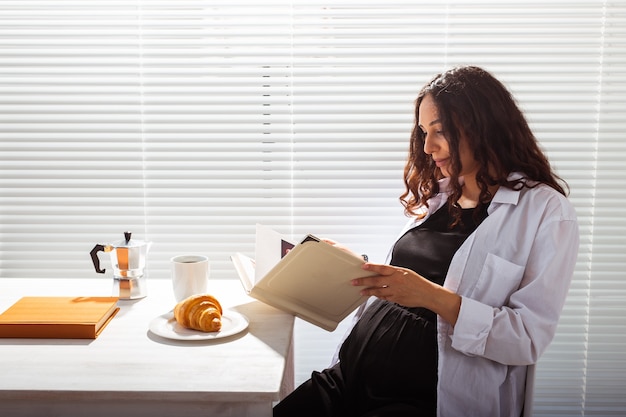 Seitenansicht der glücklichen schwangeren Frau, die Buch liest, während Morgenfrühstück mit Kaffee und