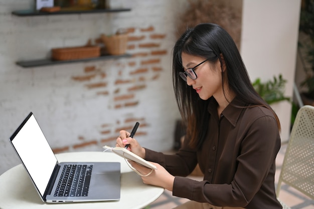 Seitenansicht der jungen Frau, die mit Zeitplanbuch und Laptop auf rundem Tisch im Café arbeitet