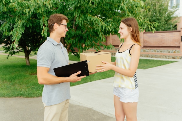 Seitenansicht der jungen glücklichen Frau, die eine Box von einem Lieferboten mit Klemmbrettern empfängt, die draußen stehen. In Freizeitkleidung gekleidet.