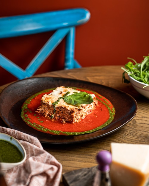 Seitenansicht der Lasagne in schwarzer Platte auf Holztisch mit blauem Stuhlhintergrund