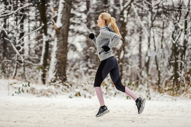 Seitenansicht der Sportlerin in Form, die am verschneiten Wintertag in der Natur joggt. Winterfitness, Übungen, kaltes Wetter