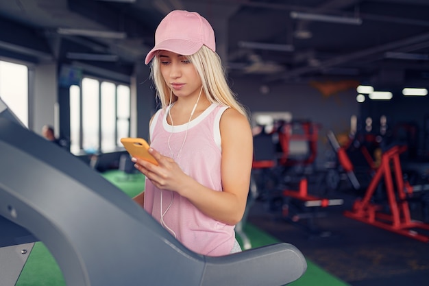 Seitenansicht der sportlichen blonden Frau in der rosa Kappe, die für das Trainieren auf dem Laufband durch Auswahl der Musik auf dem Smartphone im Fitnessstudio vorbereitet