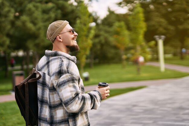 Seitenansicht des bärtigen Hipster-Studenten sieht seitlich aus, während er im Ruhetag des städtischen Stadtparks spazieren geht