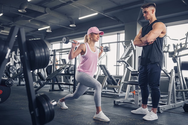 Seitenansicht des fokussierten und motivierten sportlichen jungen blonden Mädchens in der Sportbekleidung, die Beinübungen macht, während hübscher muskulöser Personal Trainer, der sie im Fitnessstudio überwacht