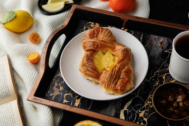 Seitenansicht des frühstückssets mit croissant auf teller und marmelade in schüssel und tasse tee auf tablett mit offenem buch aus zitronenmandarine und kleiner blume auf buch auf stoff auf schwarzem hintergrund