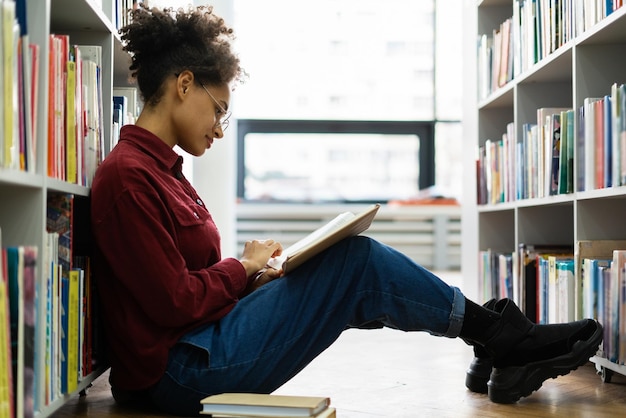 Seitenansicht des gemischtrassigen Studentenmädchens, das sich konzentriert fühlt, während es auf dem Boden sitzt und ein Buch liest Frau, die sich auf das Prüfungskonzept vorbereitet