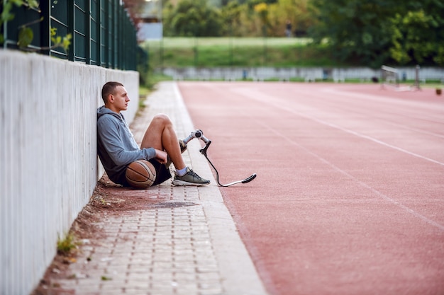 Seitenansicht des gut aussehenden ernsthaften sportlichen behinderten Mannes in Sportbekleidung und mit künstlichem Bein, das auf der Rennstrecke mit Basketballball sitzt.