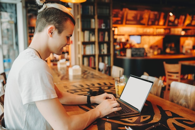 Seitenansicht des jungen Mannes mit kreativem Haarschnitt im weißen Hemd, das am Fenster am hohen Holztisch sitzt und auf Laptop-Tastatur lächelnd tippt