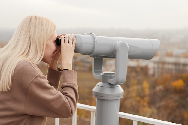 Seitenansicht des jungen weiblichen Touristen mit blondem Haar in der Sightseeing-Stadt der Freizeitkleidung mit Teleskopferngläsern, die auf Aussichtsplattform am Herbsttag stehen