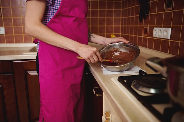 Seitenansicht des Konditors in der rosa Schürze, die geschmolzene Schokoladenmasse in der Schüssel mit einem hölzernen Spatel mischt, der in ihrer Hausküche steht