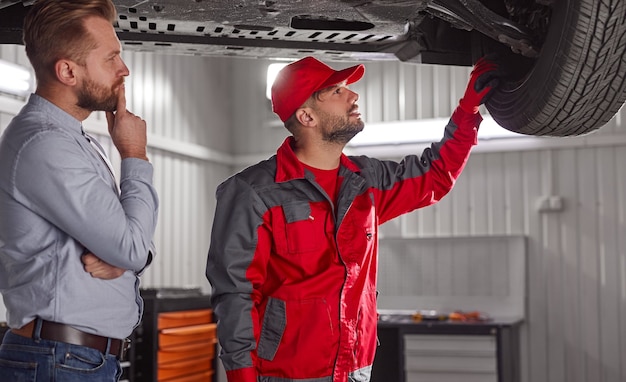 Seitenansicht des konzentrierten jungen Geschäftsmannes in der formellen Kleidung und im Mechaniker in der Uniform, die Details des gebrochenen Autos in der Garage prüft