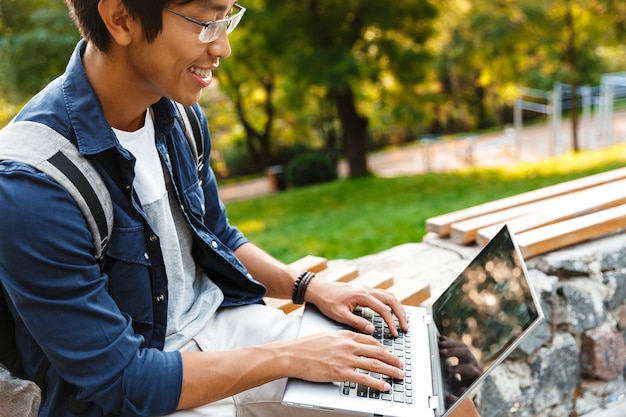 Seitenansicht des lächelnden asiatischen männlichen studenten in den brillen unter verwendung des laptop-computers beim sitzen auf der bank im park