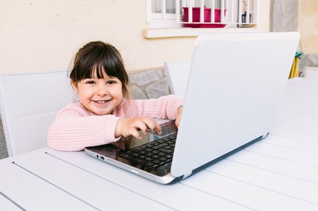 Seitenansicht des Mädchens trägt rosa Pullover, sitzt vor Laptop, auf der Terrasse, lächelnd. Online-Fernunterricht, Kindheit und Technologiekonzept.