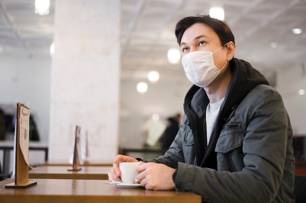 Seitenansicht des Mannes mit der medizinischen Maske, die an einem Tisch sitzt, um Kaffee zu haben