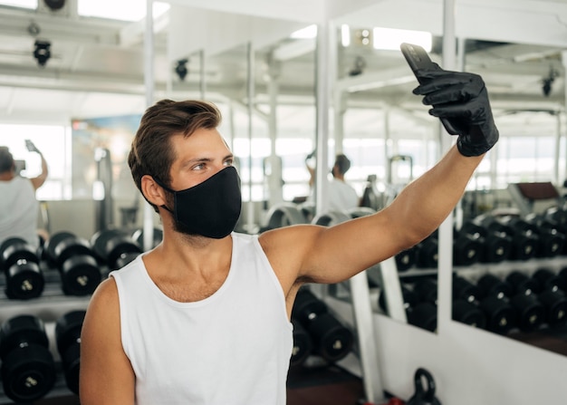 Seitenansicht des Mannes mit der medizinischen Maske, die ein Selfie an der Turnhalle nimmt