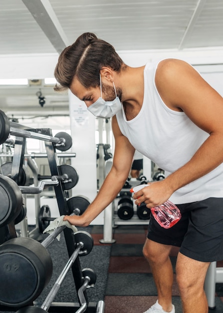 Foto seitenansicht des mannes mit der medizinischen maske, die fitnessgeräte desinfiziert