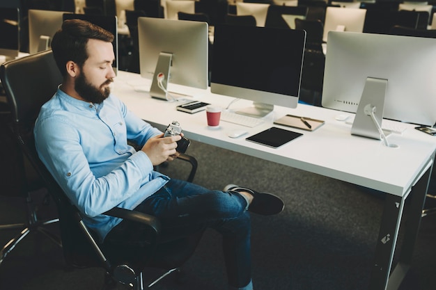 Seitenansicht des schönen jungen Mannes, der auf Stuhl sitzt und Fotos auf Kamera während der Arbeit im Büro betrachtet