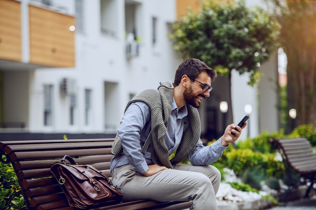 Seitenansicht des stilvollen lächelnden kaukasischen Geschäftsmannes, der auf Bank im Park sitzt und Smartphone verwendet. Neben ihm steht eine Lederhandtasche und im Hintergrund Gebäude.
