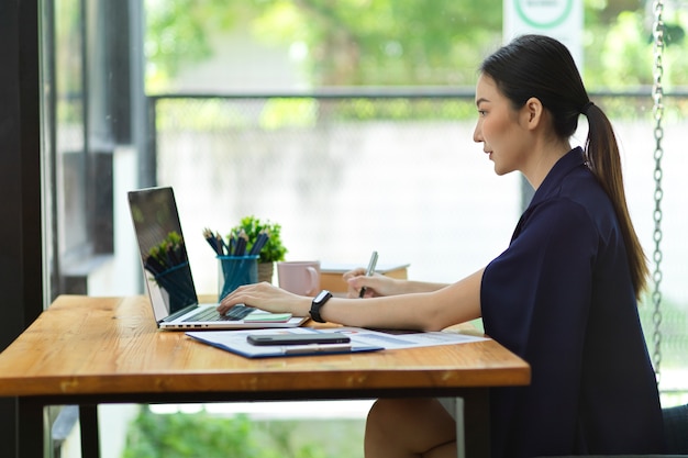 Seitenansicht einer attraktiven asiatischen Geschäftsfrau, die an einem Laptop im Arbeitsbereich im Büro arbeitet, mit verschwommener Außenansicht im Hintergrund