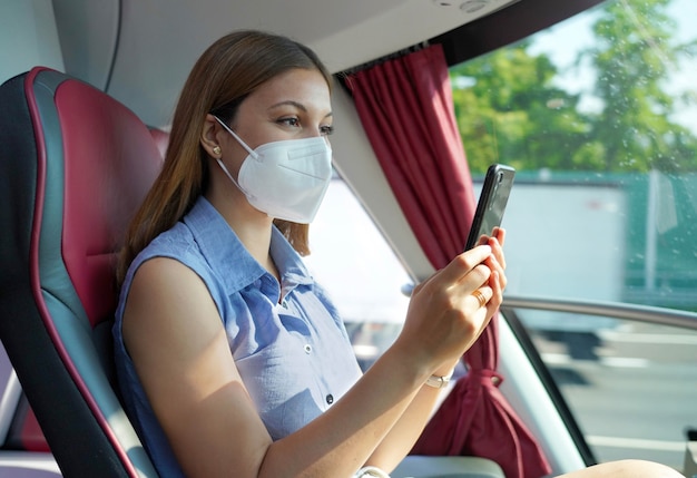 Seitenansicht einer entspannten Frau mit KN95 FFP2-Gesichtsmaske mit Smartphone-App. Buspassagier mit Schutzmaske unterwegs SMS auf dem Handy.