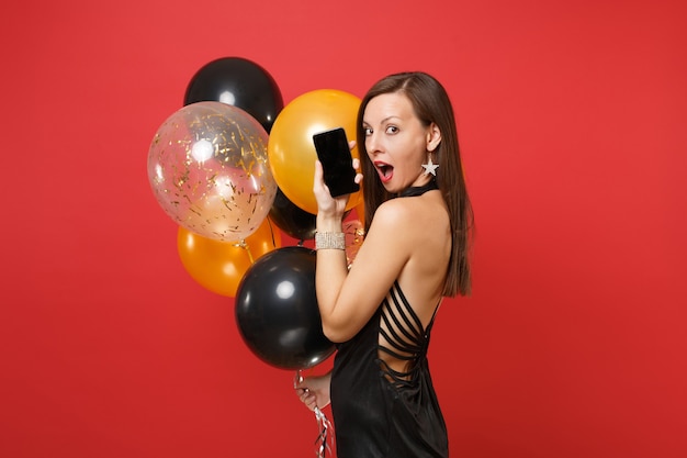 Seitenansicht einer erstaunten Frau im schwarzen Kleid, die feiert, Handy mit leerem schwarzem leerem Bildschirm hält, Luftballons einzeln auf rotem Hintergrund. Happy New Year Geburtstag Mockup Urlaub Party Konzept.