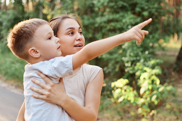 Seitenansicht einer erstaunten Mutter, die Kind in den Händen hält, kleiner Sohn, der Mutter etwas weit weg zeigt, mit dem Finger zeigt, Frau, die mit erstauntem Ausdruck in die Ferne schaut.