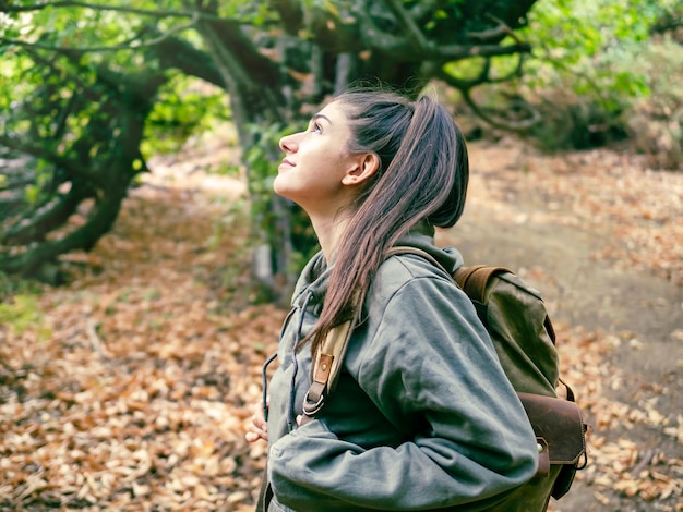 Seitenansicht einer Frau mit Rucksack im Wald, die die wilde Natur bewundert und mit einem Lächeln nach oben schaut