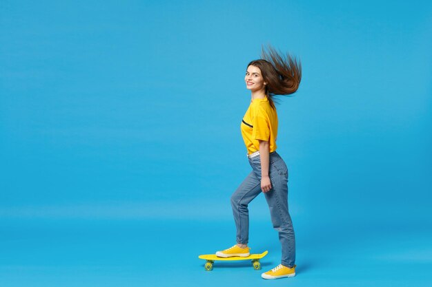 Seitenansicht einer fröhlichen jungen Frau in lebhafter Freizeitkleidung mit flatterndem Haar, die mit gelbem Skateboard isoliert auf blauem Wandhintergrund im Studio steht. People-Lifestyle-Konzept. Mock-up-Kopienbereich.