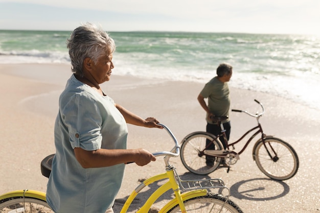 Seitenansicht einer gemischtrassigen älteren Frau und eines älteren Mannes mit Fahrrädern am Strand, die an einem sonnigen Tag wegschauen