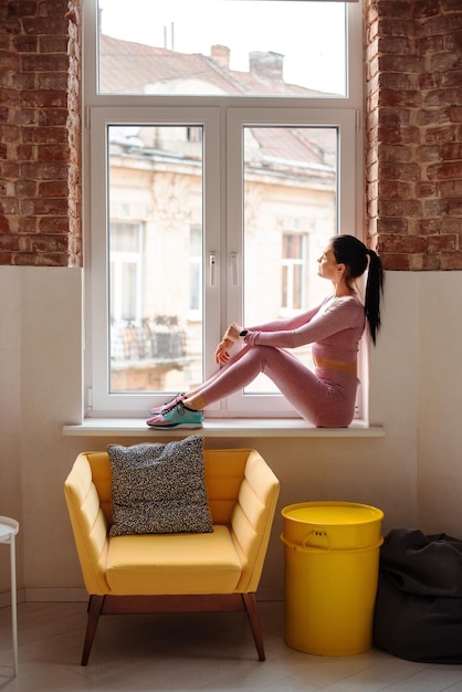 Seitenansicht einer gesunden und fitten Frau in stilvoller Sportkleidung, die auf der Fensterbank einer modernen Wohnung sitzt Junge kaukasische Dame, die sich nach dem Training zu Hause ausruht