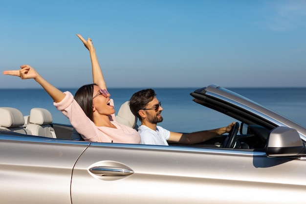 Seitenansicht einer glücklichen Frau, die die Hände hochhält und mit ihrem Freund im Cabrio sitzt