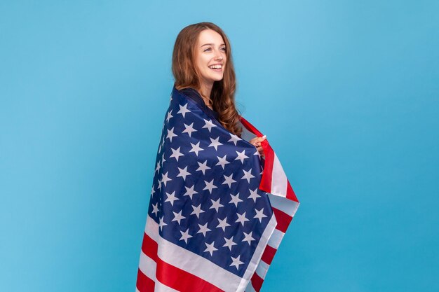 Seitenansicht einer glücklichen Frau, die einen gestreiften Pullover im lässigen Stil trägt und die Flagge der Vereinigten Staaten von Amerika hält, um die Unabhängigkeit des Arbeitstages zu feiern Studioaufnahme isoliert auf blauem Hintergrund