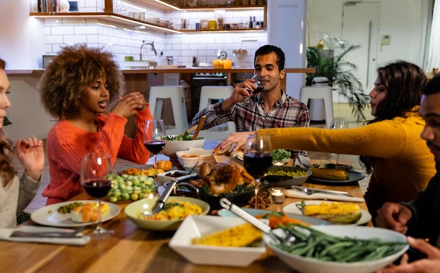 Seitenansicht einer Gruppe junger, multiethnischer, männlicher und weiblicher Freunde, die zusammen zu Hause um einen Tisch sitzen, reden, trinken und das Thanksgiving-Abendessen servieren