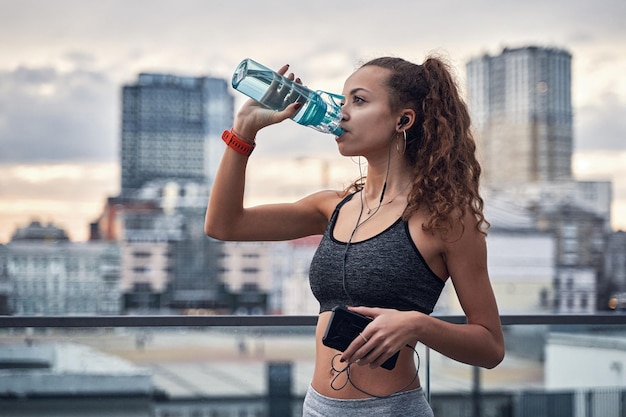 Seitenansicht einer jungen athletischen Frau, die Wasser aus einer Sportflasche trinkt, während sie Musik auf Stadthintergrund hört