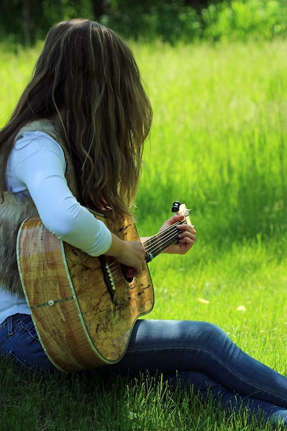 Foto seitenansicht einer jungen frau, die gitarre spielt, während sie auf einem grasbewachsenen feld im park sitzt