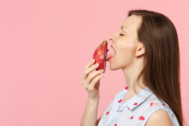 Seitenansicht einer jungen Frau in Sommerkleidung, die die Hälfte frischer reifer Pitahayas leckt, Drachenfrucht isoliert auf rosa Pastellwandhintergrund. Menschen lebendiger Lebensstil entspannen Urlaubskonzept. Mock-up-Kopienbereich.