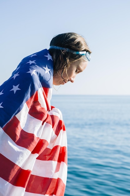 Seitenansicht einer jungen Frau mit nassen Haaren, die in amerikanischer Flagge und Schwimmbrille im Sonnenlicht nach unten schautSeascape im Hintergrund