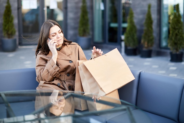 Seitenansicht einer jungen Frau mit Sitzen auf einer Holzbank, Papiertüten mit Einkäufen in der Nähe, verwendet das Mobiltelefon, scrollt durch neue Nachrichten, sucht nach Informationen über das Einkaufszentrum