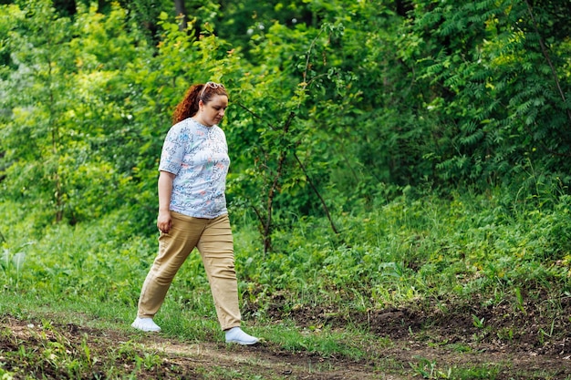 Seitenansicht einer jungen, prallen, drallen Frau mit langen, lockigen roten Haaren, die auf dem Weg im Parkwald zwischen grünen Bäumen spazieren geht Sommer