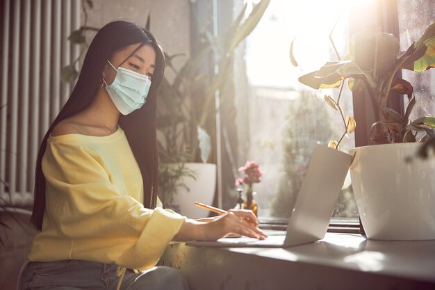 Seitenansicht einer jungen schönen asiatischen Dame, die eine medizinische Maske trägt, wenn sie während der Quarantäne an einem Laptop im Café arbeitet