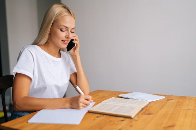 Seitenansicht einer lächelnden blonden jungen Frau, die lernt, sich Notizen zu machen, einen Aufsatz in ein Notizbuch zu schreiben und auf dem Smartphone zu sprechen, während sie akademische Forschung zur Vorbereitung auf die Prüfung durchführt