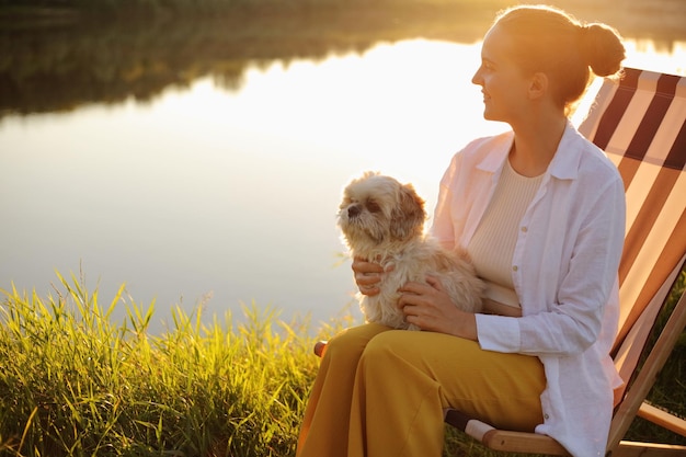 Seitenansicht einer lächelnden, träumenden jungen erwachsenen Frau mit weißem Hemd und ihrem pekinesischen Hund, die bei Sonnenuntergang in der Nähe des Flusses sitzt und auf das Wasser blickt und die Aussicht genießt