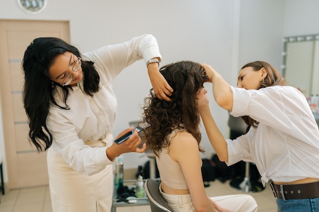 Seitenansicht einer lockigen jungen Frau, die in einem Schönheitssalon Augenschatten auftut Friseur, der lange Haare einer jungen Frau mit einer Haarbürste kämmt